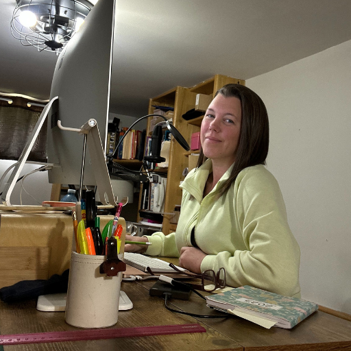 A woman wearing a yellow zip up shirt holding a green pencil standing at her desk in front of a large computer screen smiling. On the desk is a closed notebook, eye glasses, a cup of colourful highlighters and a pink ruler in front of the cup.