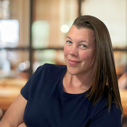 A woman with long brown hair standing in front of a blurred background leaning to the right looking at the camera. She is wearing a dark blue t-shirt.