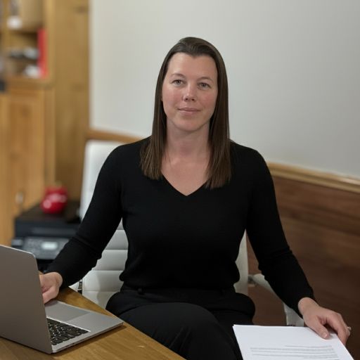 A woman wearing a yellow zip up shirt holding a green pencil standing at her desk in front of a large computer screen smiling. On the desk is a closed notebook, eye glasses, a cup of colourful highlighters and a pink ruler in front of the cup.