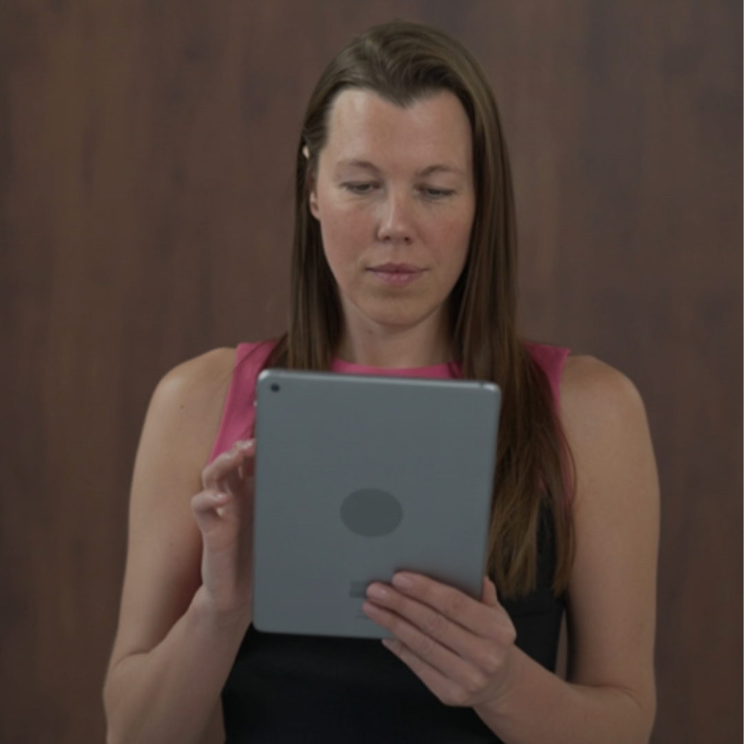 A woman with long brown hair standing in front of a blurred background leaning to the right looking at the camera. She is wearing a dark blue t-shirt.